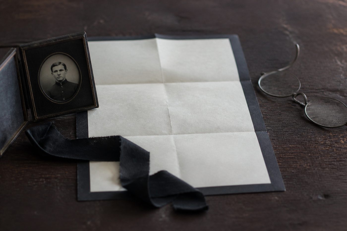 A black bordered letter unfolded on a dark wooden table beside a small cased photograph of a young man, a length of black crape ribbon and a pair of reading spectacles.